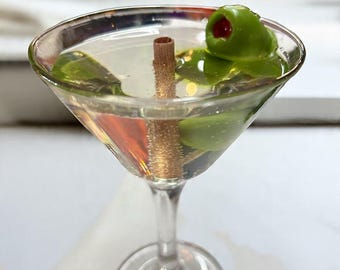 May include: A clear martini glass filled with a pale liquid, green olives, and a cinnamon stick. The martini glass sits on a white surface. The olives have a red center. The drink is a classic cocktail.