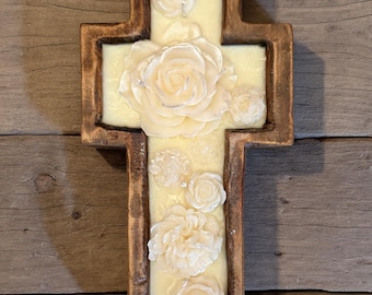 May include: Wooden cross with a cream-colored center and carved white floral accents. The cross has a rustic, brown frame and is set against a wooden background. The flowers include roses and other detailed blossoms.