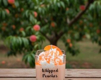 May include: A square glass candle with "Whipped Peaches" text, topped with white and orange wax, and a peach slice. The candle sits on a wooden surface with a blurred peach tree background.