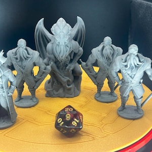 May include: A collection of gray miniature figurines, including a central winged creature and several humanoid figures with tentacled faces. A multi-sided die with visible numbers sits on a yellow surface.
