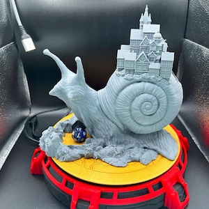 May include: A gray 3D-printed sculpture of a snail carrying a miniature town on its back. The snail sits on a yellow and red platform, with a blue and white die nearby. The sculpture is detailed and whimsical.