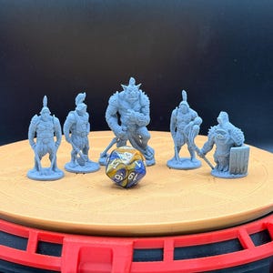 May include: A collection of gray miniature fantasy figures, including warriors and a large orc, arranged around a multi-sided die. The figures are on a tan circular platform with a red base, suitable for tabletop gaming.