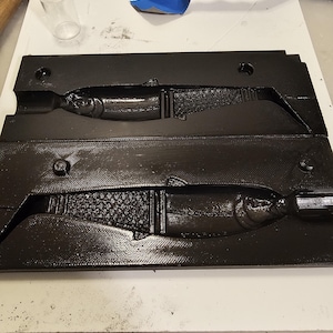 May include: A black plastic mould for making fishing lures. The mould has two cavities, each shaped like a fish. The mould is sitting on a white surface.