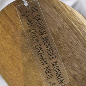 Könnte beinhalten: Ein rechteckiges Lesezeichen aus klarem Acryl mit dem schwarz aufgedruckten Text "DIE GRÖSSTEN ABENTEUER BEGINNEN MIT EINEM EINZIGEN BUCH!" und kleinen rautenförmigen Symbolen. Eine silberne Kordel ist durch ein Loch am oberen Ende befestigt.