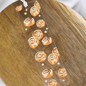 Könnte beinhalten: Ein rechteckiges Lesezeichen aus transparentem Acryl mit einem wiederkehrenden Muster aus illustrierten Zimtschnecken mit weißem Zuckerguss. Das Design enthält kleine beige Punkte und den braunen Schriftzug "COZY BAKERY" sowie eine weiße Kordel.