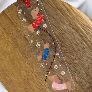 Könnte beinhalten: Ein transparentes, rechteckiges Lesezeichen mit drei braunen Dackeln, die rote, blaue und rosafarbene Pullover tragen. Die Hunde sind in eine bunte Lichterkette verwickelt, umgeben von weißen Schneeflockenmotiven und einer roten Kordel oben.