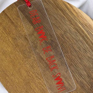 Könnte beinhalten: Ein rechteckiges Lesezeichen aus transparentem Acryl mit dem vertikal aufgedruckten roten Text "DEAR BVNK, BE BACK SVVN!". Das Lesezeichen hat oben ein kleines Loch, durch das eine dünne rote Kordel gezogen ist, die eine Schlaufe bildet.