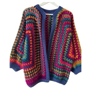 Könnte beinhalten: Ein farbenfroher, offener Häkelcardigan im Granny-Square-Muster. Der Pullover hat eine lebendige Mischung aus Farben, darunter Blau, Rosa, Orange und Türkis. Die Kanten und der Kragen sind einfarbig blau.