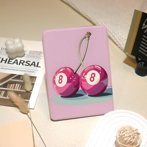 May include: A pink tablet case featuring a graphic of two cherries, each resembling an eight-ball with the number 8. The cherries have a green stem and are set against a light pink background. The case is rectangular.