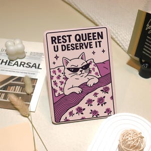Könnte beinhalten: Eine rosa rechteckige Hülle mit einer Katze, die eine Sonnenbrille trägt und in einem Bett ruht. Der Text "REST QUEEN U DESERVE IT" steht oben. Das Bett hat ein Blumenmuster und die Katze ist von Sternen umgeben.