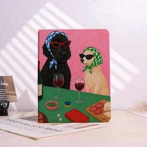 May include: A notebook featuring an illustration of two dogs, one black and one beige, wearing sunglasses and headscarves. They are depicted with wine and snacks on a green surface against a pink backdrop.