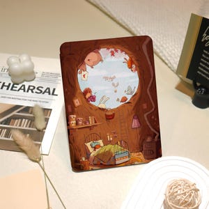 May include: A brown and tan illustrated case for an e-reader. The case features a whimsical scene of a tree with a hole in it. Inside the tree, a small animal is sleeping in a bed. Outside the tree, other small animals are flying in the sky. The text "The End" is visible in the image.