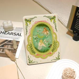 May include: A cream-coloured book cover featuring an illustration of two mermaids in a green pond. The design is framed by a green and pink border with floral accents. The cover has a vintage aesthetic.