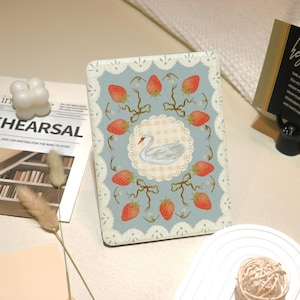 May include: A rectangular, light blue case with a strawberry and swan design. The case features a white lace border and a central image of a swan surrounded by strawberries and bows. The case is likely for a tablet or e-reader.