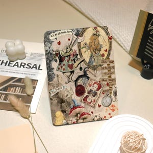 Könnte beinhalten: Eine Illustration im Vintage-Stil mit Figuren aus Alice im Wunderland, darunter der Hutmacher, die Herzkönigin und Alice selbst. Die Illustration zeigt ein fantasievolles und farbenfrohes Design mit Text, der "We're all mad here", "This Way NOWHERE", "That Way" und "Drink Me" lautet.