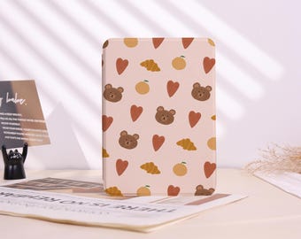 May include: A rectangular case featuring a repeating pattern of brown bear faces, red hearts, yellow croissants, and orange fruit on a light pink background. The case is likely for a tablet or e-reader.