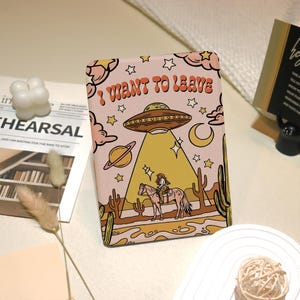 May include: A rectangular case with a pink background featuring a retro illustration. The design includes a UFO, a cowboy on a horse, cacti, stars, and the text "I WANT TO LEAVE".
