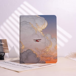 May include: A rectangular notebook featuring a printed illustration of a small red biplane flying through large, voluminous clouds against a sunset sky with shades of blue, grey, and orange. The cover has a smooth finish and rounded corners.