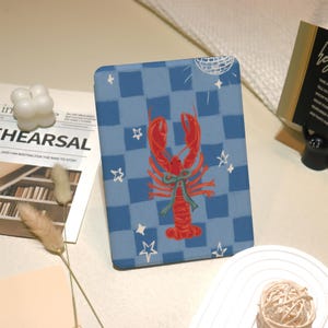 May include: A blue and white checkered e-reader cover with a red lobster illustration and a disco ball. The lobster is wearing a green bow.