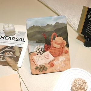 May include: A decorative tablet case featuring a picnic scene with a basket, hat, wine, and an open book with a heart. The background shows a lake and mountains, with a soft color palette.