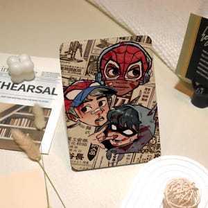 May include: A colourful illustration of three superheroes, including Spider-Man, with a newspaper background. The superheroes are drawn in a cartoon style with exaggerated features and are wearing masks. The image is set against a backdrop of a newspaper with Japanese text.