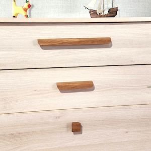 May include: A light-colored wooden dresser with three drawers, each featuring a natural wood handle. The top of the dresser has decorative items, including a small sailboat and a yellow and red animal figurine.
