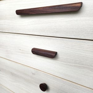Op de afbeelding: Close-up van een lichtgekleurde houten dressoir met donkerbruine houten ladehandgrepen. De bovenste lade heeft een lange, gebogen handgreep, terwijl de andere laden kortere, rechthoekige handgrepen en een ronde knop hebben. De houtnerf is zichtbaar.