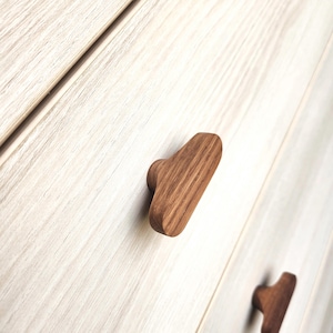 May include: Close-up of wooden cabinet knobs. The knobs are a warm brown color and have a rectangular shape with a rounded end. The wood grain is visible. The knobs are attached to a light-colored wooden cabinet.