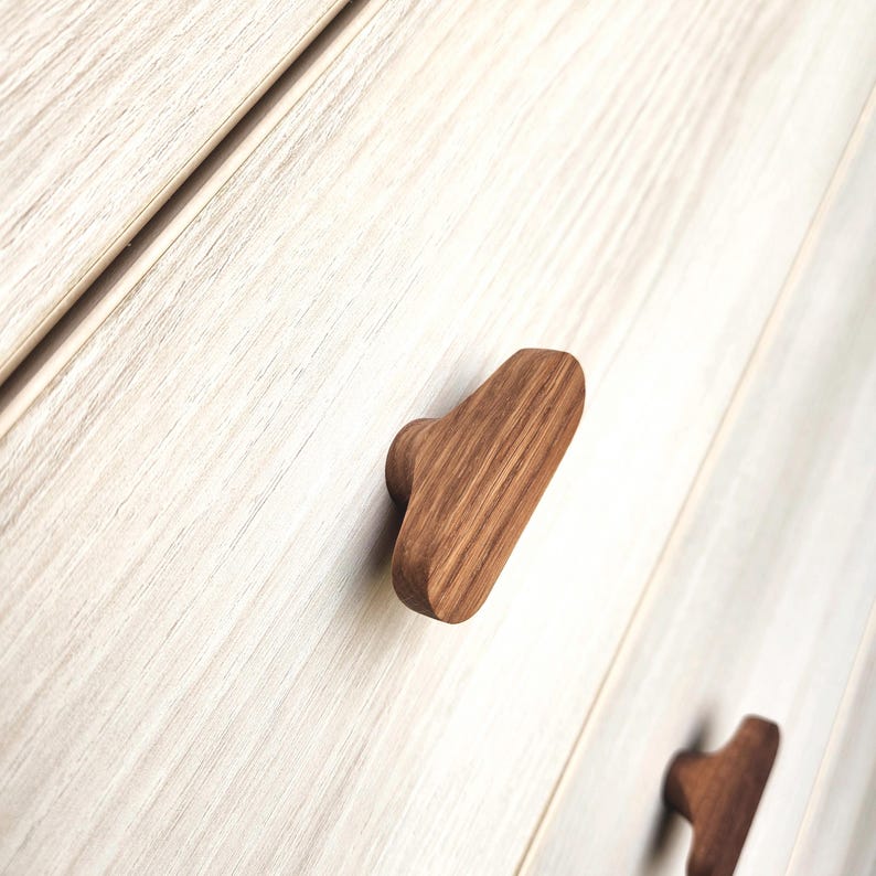 May include: Close-up of wooden cabinet knobs. The knobs are a warm brown color and have a rectangular shape with a rounded end. The wood grain is visible. The knobs are attached to a light-colored wooden cabinet.