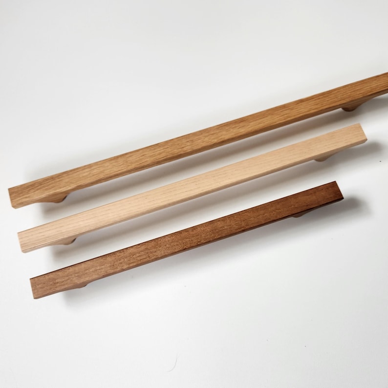 May include: Three wooden cabinet pulls in varying shades of brown. The top pull is a light brown, the middle is a natural wood tone, and the bottom is a dark brown. Each pull has a rectangular shape with a curved handle.