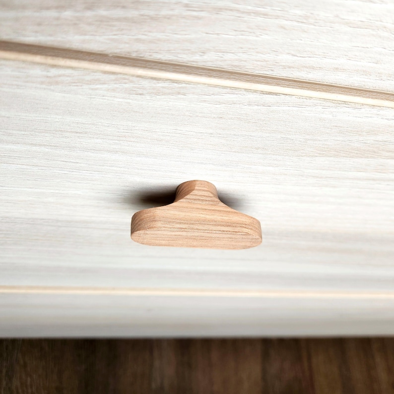 May include: A close-up of a wooden cabinet knob with a rounded top and a flat, rectangular base. The knob is made of light-colored wood and is mounted on a white cabinet door with a wood grain pattern.