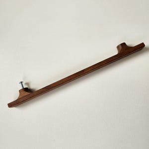 May include: A long, dark brown wooden cabinet pull with a curved handle and a flat, rectangular body. A silver screw is attached to one end. The pull is angled across a neutral background.