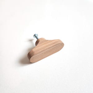 May include: A wooden cabinet knob with a light brown finish. The knob is shaped like a small, stylized coat hanger and features a silver screw for attachment. The wood grain is visible, adding texture to the simple design.