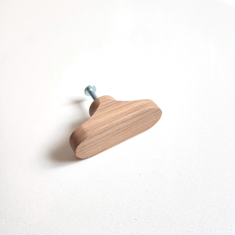 May include: A wooden cabinet knob with a light brown finish. The knob is shaped like a small, stylized coat hanger and features a silver screw for attachment. The wood grain is visible, adding texture to the simple design.