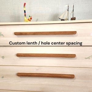 Puede incluir: Primer plano de una cómoda de madera blanca con tres tiradores horizontales de madera marrón. El texto "Custom length / hole center spacing" está impreso en negro. Una colorida figura de gato y un pequeño velero están encima.