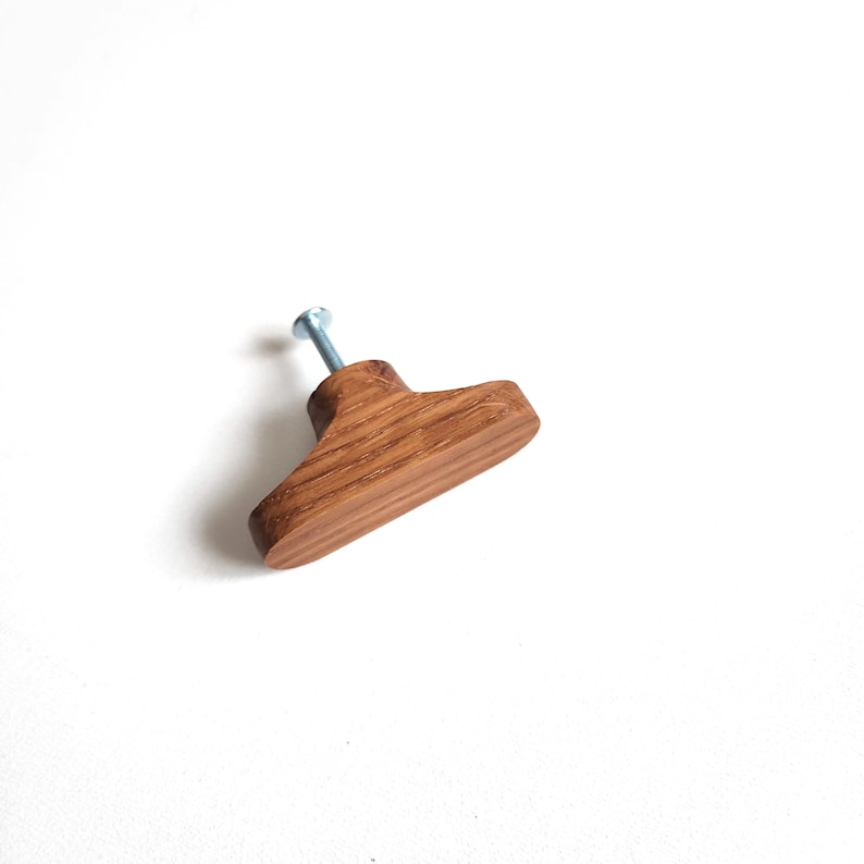 May include: A wooden cabinet knob with a T-shape design. The knob is made of light brown wood and has a silver screw attached. The wood grain is visible. The knob is designed for furniture.
