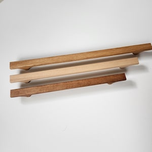 May include: Three wooden cabinet pulls in varying shades of brown. The top pull is a light oak color, the middle pull is a natural wood tone, and the bottom pull is a dark walnut color. Each pull has a rectangular shape with rounded ends.