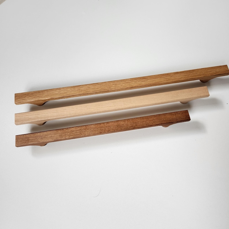 May include: Three wooden cabinet pulls in varying shades of brown. The top pull is a light oak color, the middle pull is a natural wood tone, and the bottom pull is a dark walnut color. Each pull has a rectangular shape with rounded ends.