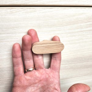 May include: A light-colored, wooden cabinet pull with an oval shape. The pull is held in a hand, showcasing its size and grain. The pull is designed for use on drawers or cabinets.