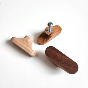 May include: Three wooden cabinet knobs in varying shapes and wood tones. One is a light brown with a curved shape, another is a darker brown oval, and the third is a light brown with a screw attached. These knobs are for furniture.