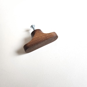 May include: A wooden cabinet knob with a dark brown finish. The knob has a rounded, rectangular shape with a metal screw attached. The hardware is designed for use on drawers or cabinet doors.