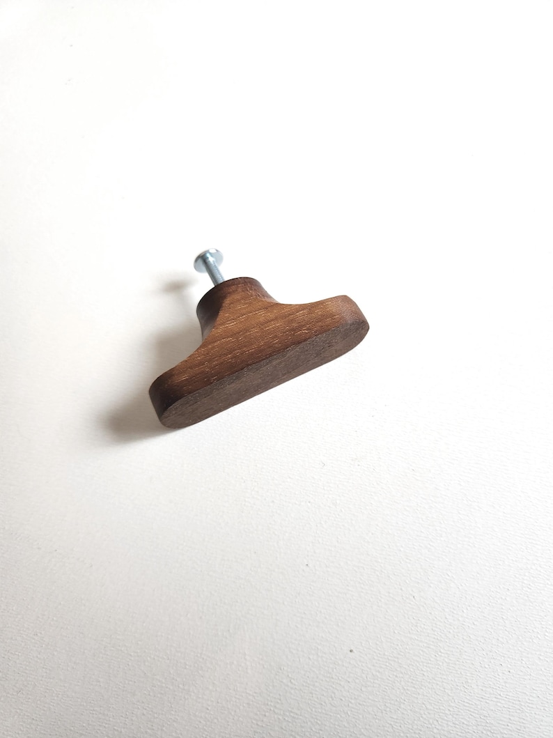 May include: A wooden cabinet knob with a dark brown finish. The knob has a rounded, rectangular shape with a metal screw attached. The hardware is designed for use on drawers or cabinet doors.