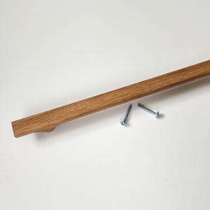 May include: A wooden cabinet pull with a natural finish, featuring a curved handle design. Two silver screws are included. The pull is made of solid wood and is designed for easy installation.