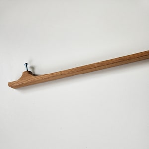 May include: A wooden wall-mounted coat rack with a natural finish. The rack is attached to the wall with a single blue screw. The design features a flat, rectangular base with a curved end, and a long, straight bar extending from it.