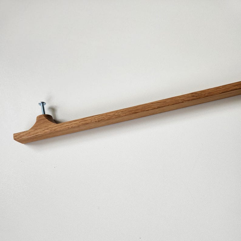 May include: A wooden wall-mounted coat rack with a natural finish. The rack is attached to the wall with a single blue screw. The design features a flat, rectangular base with a curved end, and a long, straight bar extending from it.