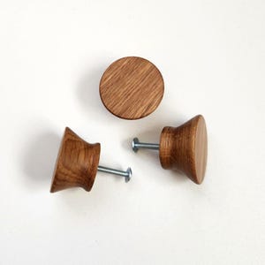 Peut inclure: Trois boutons de meuble en bois avec un fini grain de bois naturel. Deux boutons sont inclinés, montrant une base évasée et un dessus rond, avec une vis argentée attachée. Le troisième bouton est un simple disque rond. Le tout sur fond blanc.
