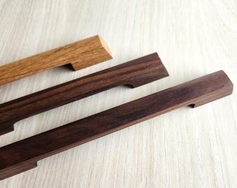 Könnte beinhalten: Nahaufnahme von drei Holzschrankgriffen in verschiedenen Brauntönen. Die Griffe sind rechteckig mit einer gebogenen Vertiefung zum Greifen. Die Holzmaserung ist sichtbar, auf einer hellen Holzoberfläche.