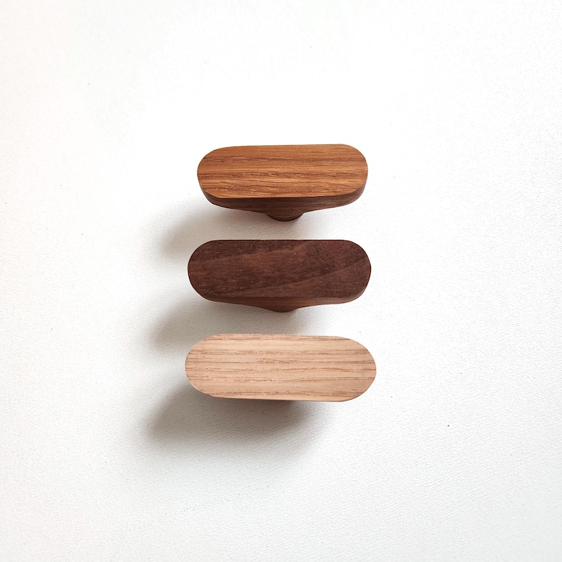 May include: Three wooden cabinet knobs in varying shades of brown. The knobs are oval-shaped with a curved base for easy gripping. The wood grains are visible, showcasing the natural textures of the materials. The knobs are likely for furniture or decorative purposes.