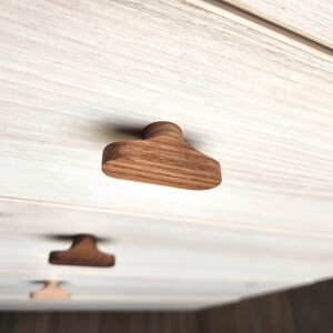May include: Close-up of wooden cabinet knobs. The knobs are a warm brown color and have a unique, geometric shape. The wood grain is visible, adding texture. The knobs are attached to a light-colored cabinet door.