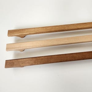 May include: Three wooden poster hangers in varying shades of brown. The top hanger is light brown, the middle is a natural wood color, and the bottom is a dark brown. Each has a curved handle-like design.